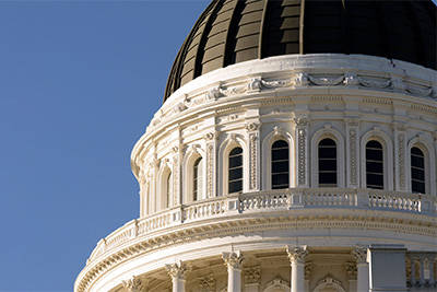 A close-up of the California State Capitol's neoclassical dome in Sacramento, showcasing its intricate white architectural details with columns, railings, and moldings against a clear blue sky.