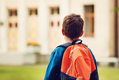 Rear view of a young boy with a orange backpack, looking towards a school building in the distance. The focus is on the child with a soft, blurred background, capturing a moment of anticipation or contemplation before entering the school.