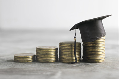 A miniature graduation cap perched on top of a stack of increasing coins against a grey background, symbolizing the cost of education and the investment in academic achievement.