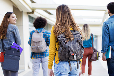 A group of diverse high school students with backpacks walking along an outdoor corridor, viewed from behind. They appear to be engaged in conversation and moving between classes, with the focus on the central student wearing a grey backpack.